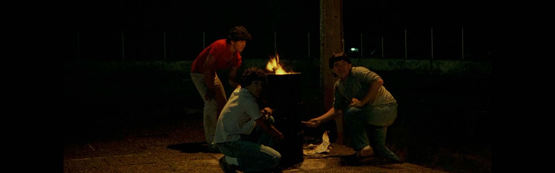 Fotograma de Los habitantes de la noche. 3 amigos adolescentes están bajo una farola, en medio de la noche, golpeando una caneca en cuyo interior resplandece fuego. 