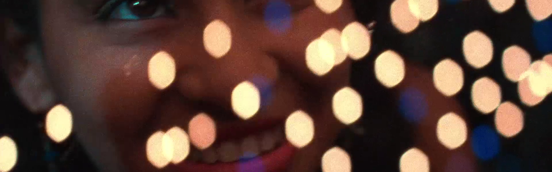 Fotograma de La vendedora de rosas. Una niña de 12 años sonríe tras una ventana, en el reflejo de la ventana se ven unas luces de fuegos artificiales.